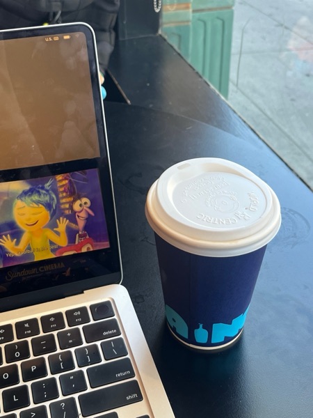 A Blue Bottle coffee cup beside a laptop playing Inside Out at a café window