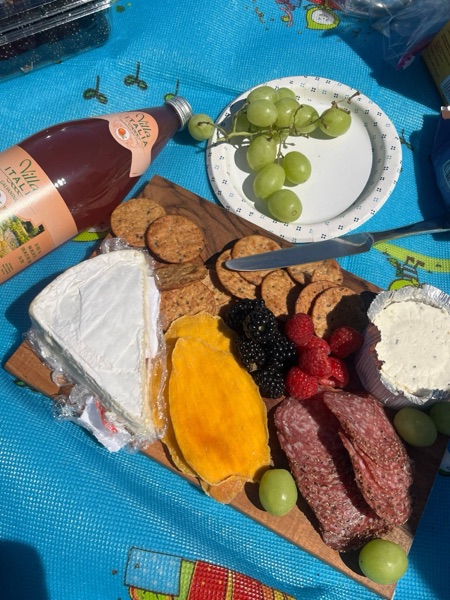A picnic board of brie, crackers, berries, dried mango, and salami on a blue mat, with green grapes and a bottle of Italian soda